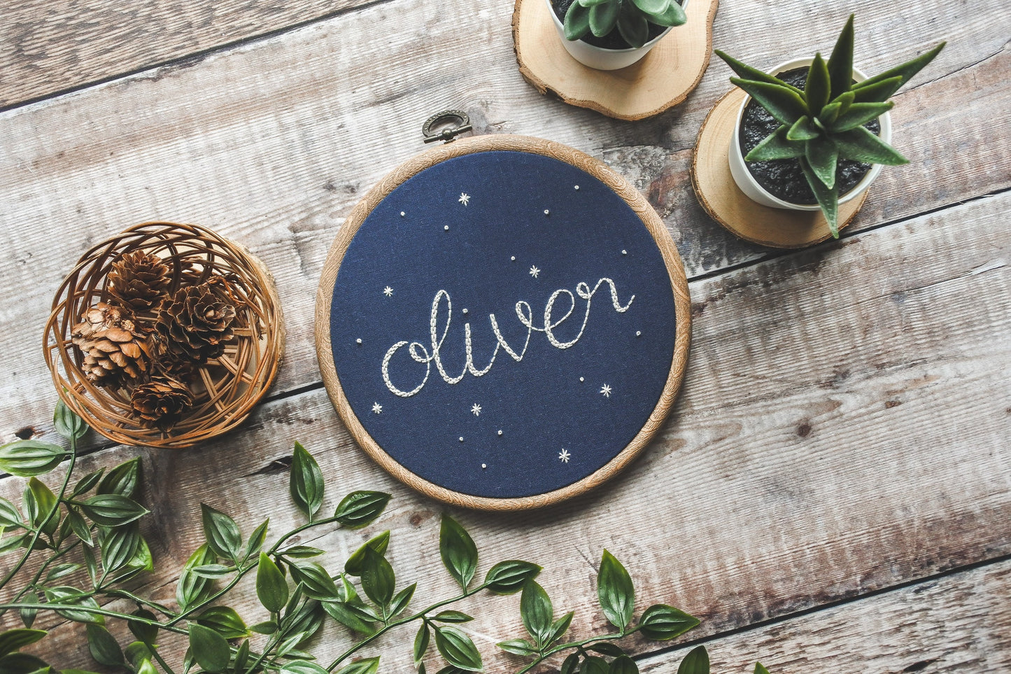 Angled view of starry name hoop with 'Oliver' embroidered in white thread on navy blue fabric, surrounded by stars