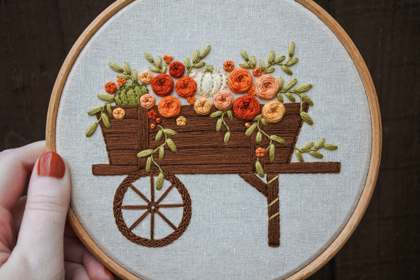 Hand holding the finished autumn wagon embroidery hoop, showcasing the completed floral design