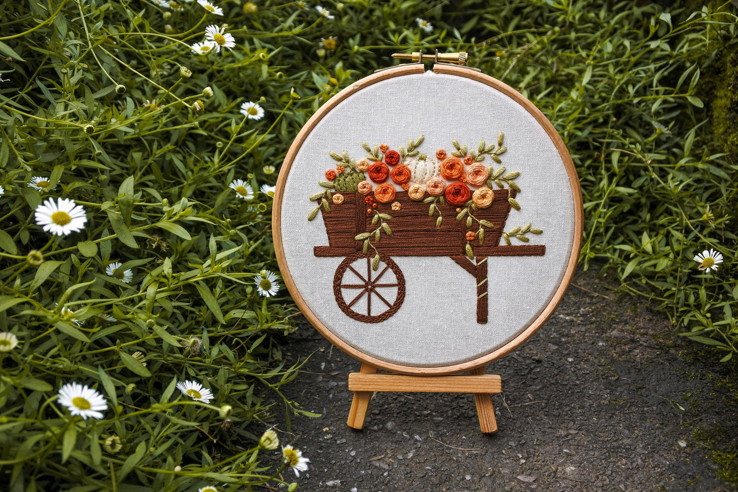 Hand-embroidered autumn wagon hoop displayed on a mini wooden easel surrounded by daisy plants