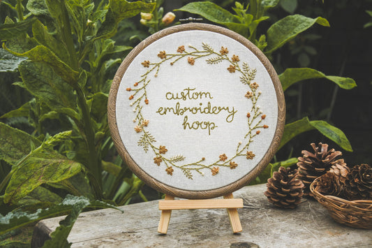 Custom autumn wreath embroidery hoop displayed on mini wooden easel, surrounded by greenery and pinecones