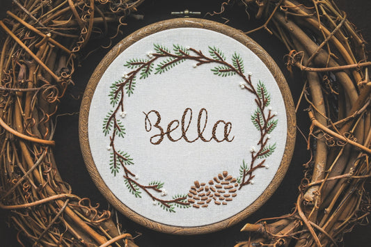 Hand embroidered winter wreath hoop with the name 'Bella', featuring pinecones, fir branches, and white berries