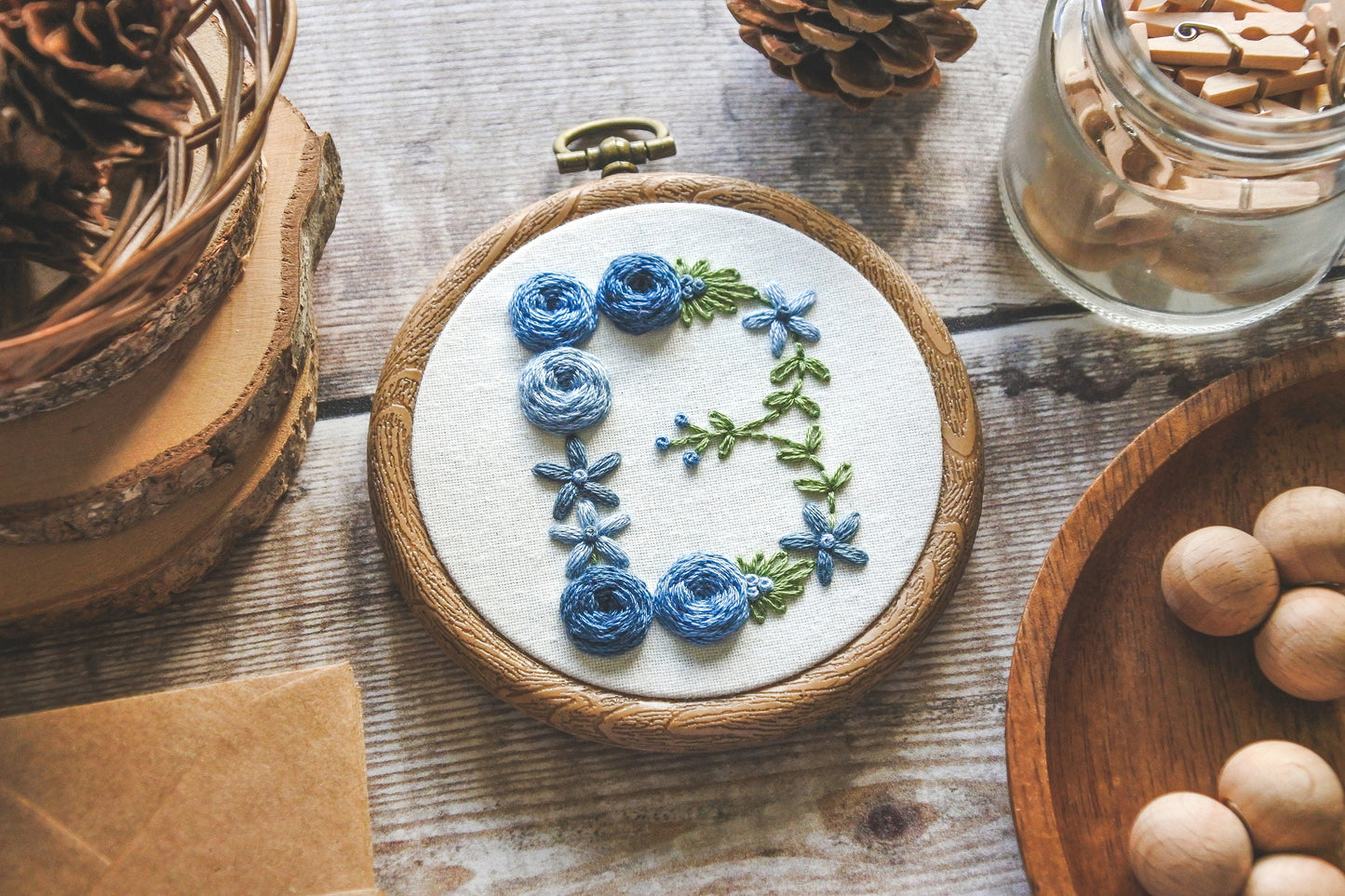 Blue floral 'B' hand-embroidered hoop with green foliage, displayed at a slight angle.
