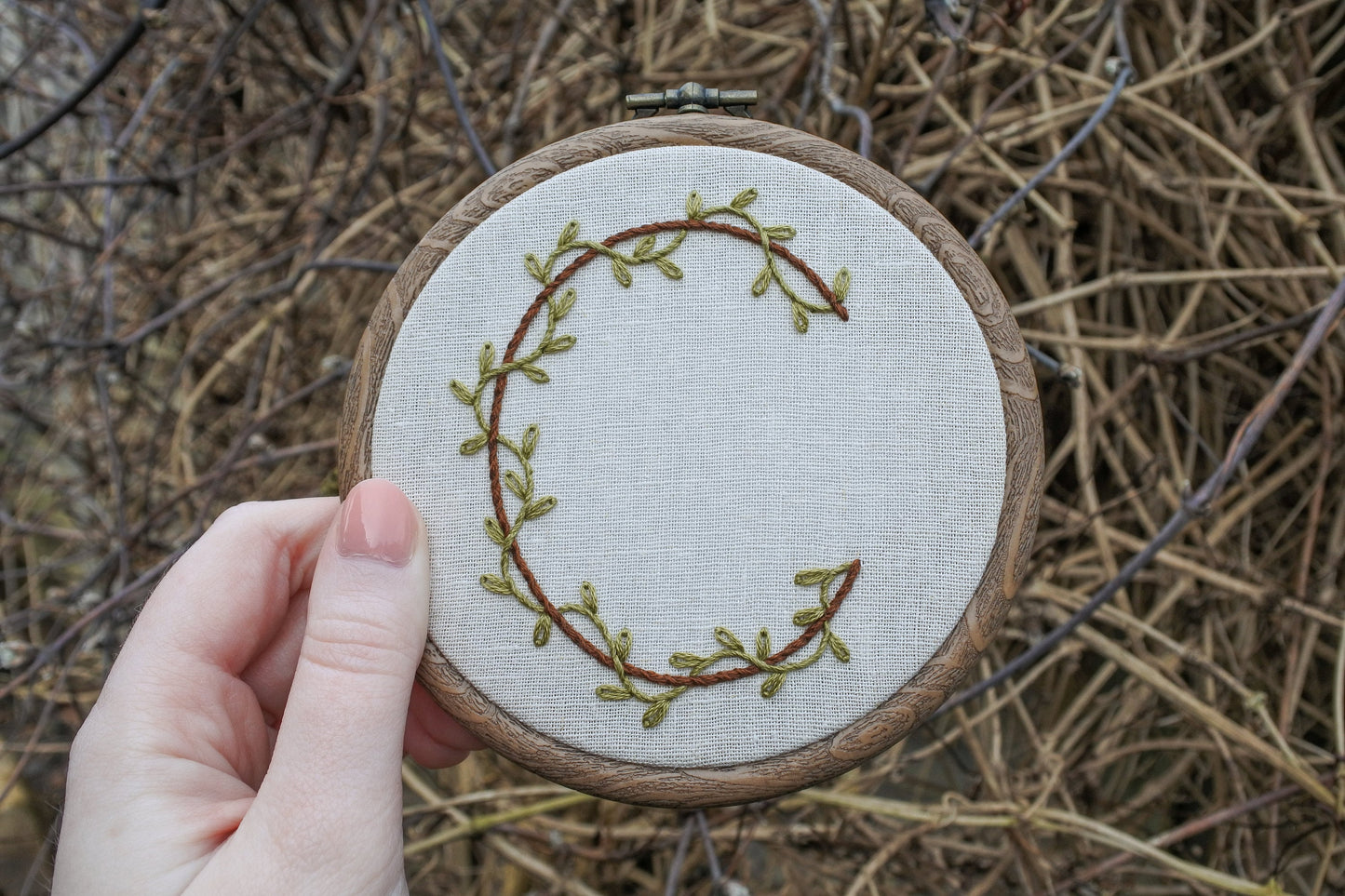 Hand holding hand embroidered letter C hoop featuring leafy green vines wrapping a round the brown letter C in a wood effect hoop frame.