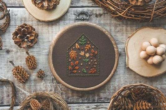 A 4-inch embroidery hoop featuring a house outline filled with warm autumn-coloured floral and botanical stitching on dark brown fabric. The hoop has a wood-effect finish.