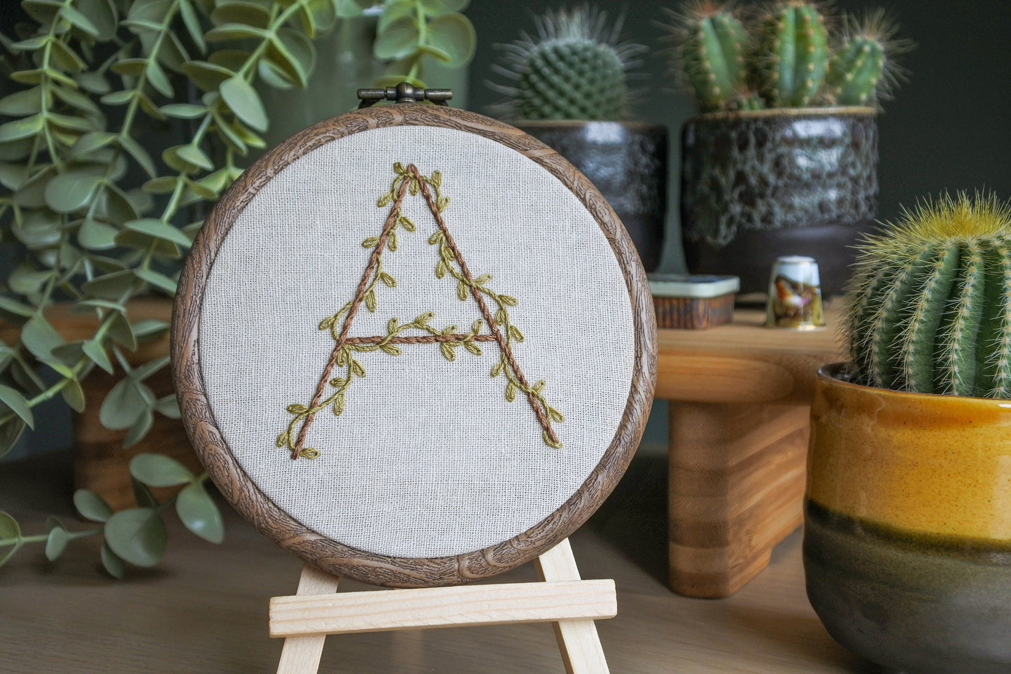 Botanical letter 'A' embroidery hoop displayed on a mini wooden easel, highlighting leafy vine details on neutral fabric.