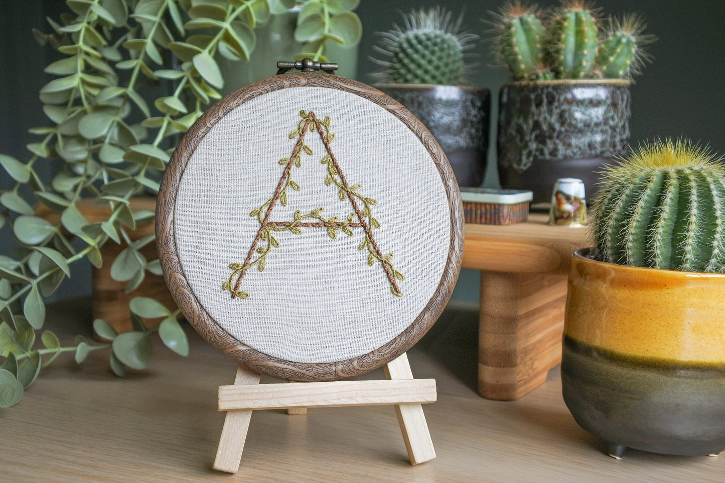 Hand-embroidered botanical letter 'A' hoop displayed on a mini wooden easel, with olive green vines and light brown stitching on neutral fabric.