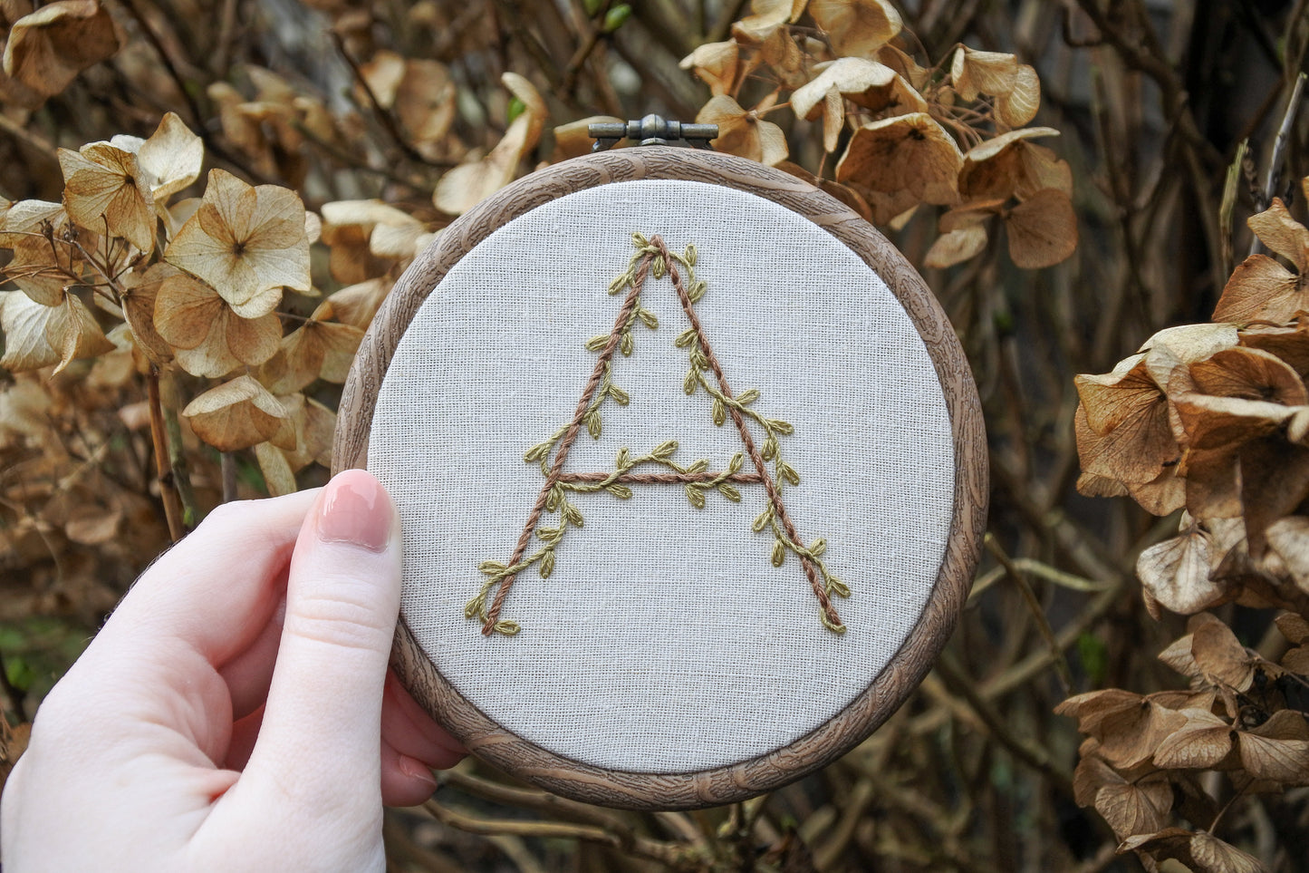 Hand holding the hand-embroidered botanical letter 'A' hoop, featuring olive green leafy vines and light brown thread.