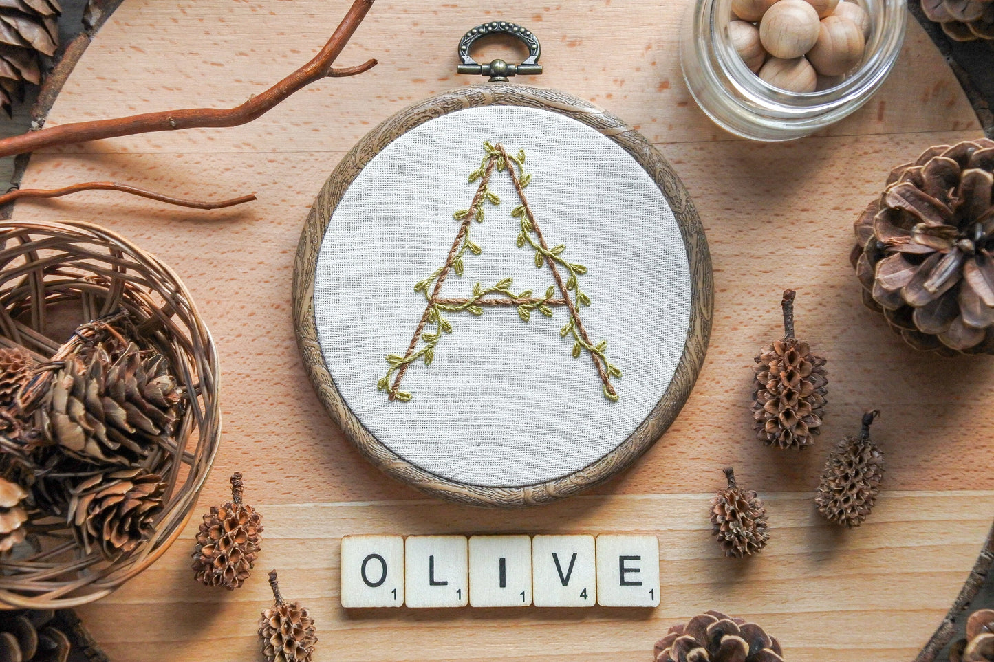 Botanical letter 'A' embroidery hoop displayed on neutral fabric with wooden Scrabble letters spelling "olive" underneath.