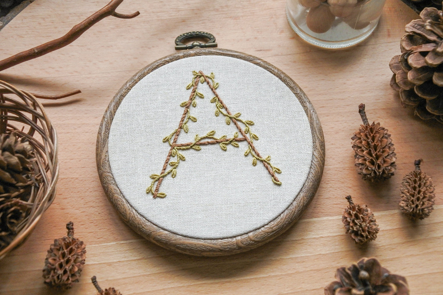 Slightly angled view of the hand-embroidered botanical letter 'A' hoop with olive green vines and light brown embroidery, set in a 4-inch wood-effect hoop.