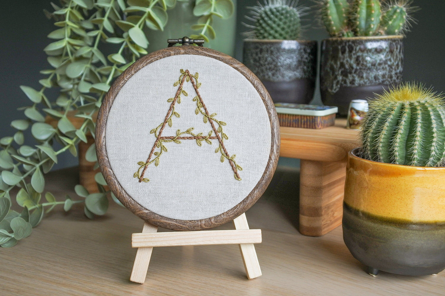Hand-embroidered botanical letter 'A' hoop displayed on a mini wooden easel, with olive green vines around the letter 'A' in light brown thread.