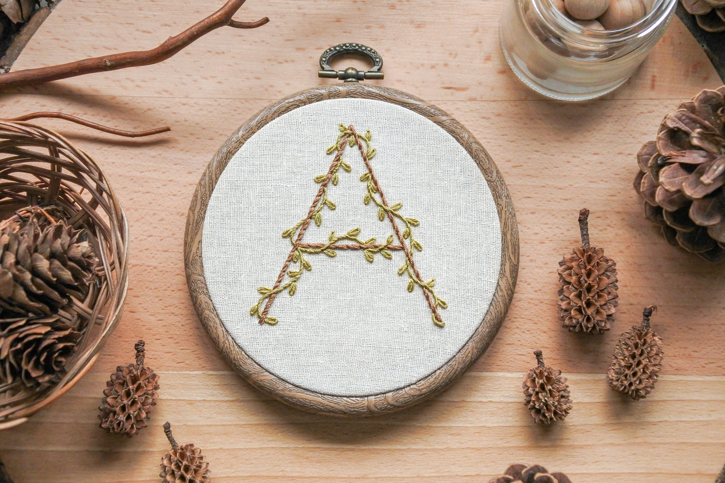 Hand-embroidered botanical letter 'A' hoop with olive green vines and light brown embroidery, set in a 4-inch wood-effect hoop.
