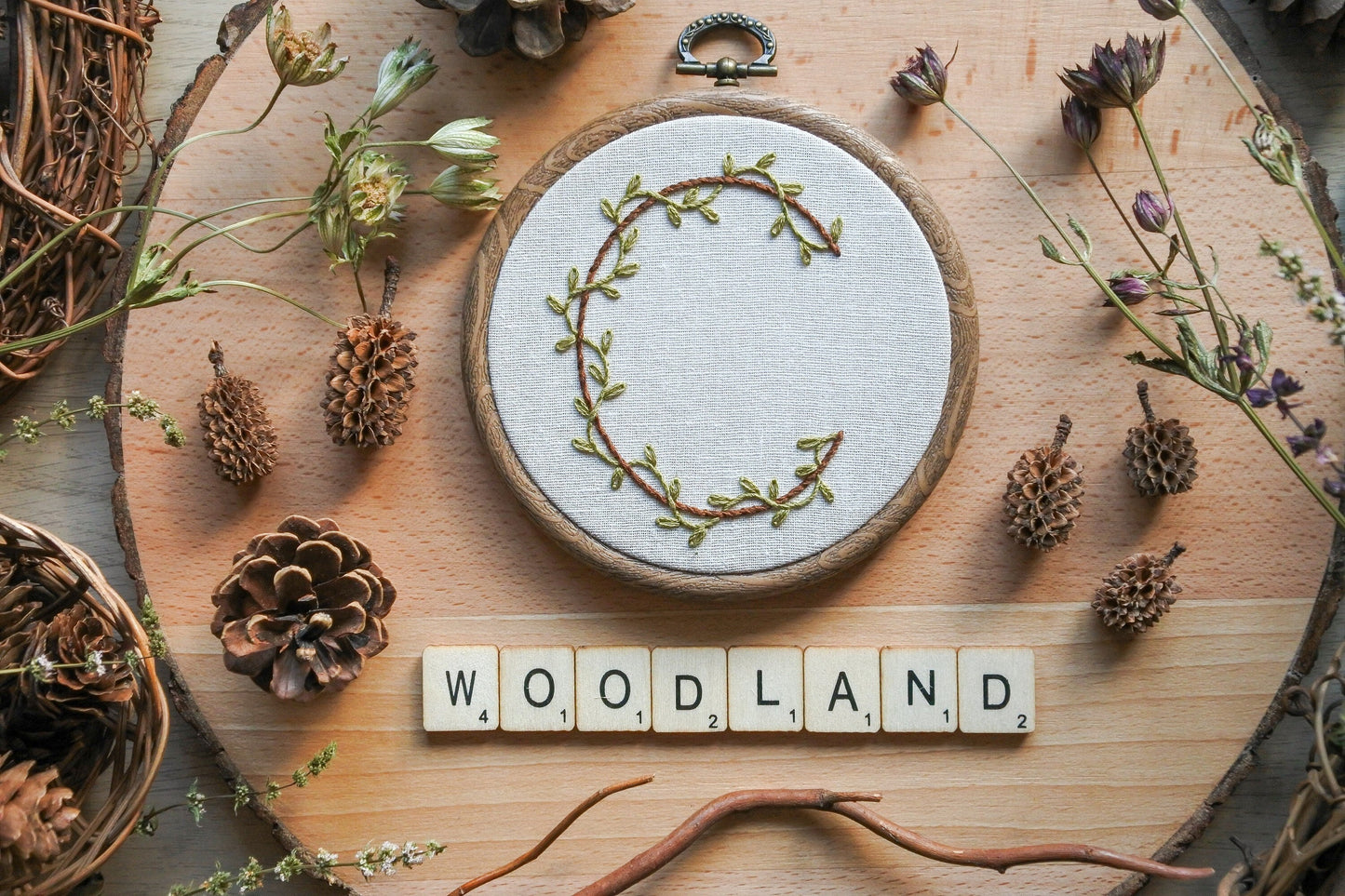 Botanical letter C embroidery hoop with green vines and brown stitched letter, displayed with wooden Scrabble tiles spelling 'woodland'