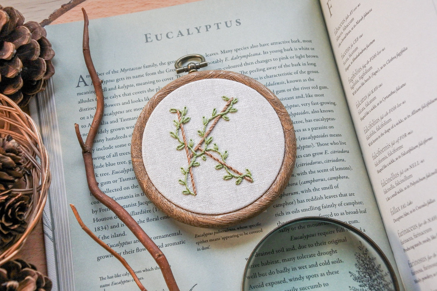 Angled view of botanical letter K embroidery hoop with eucalyptus green vines and pale brown stitched letter on neutral fabric in a 3-inch wood-effect hoop