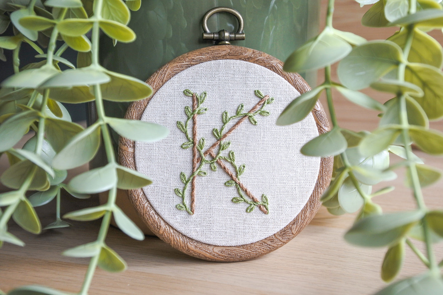 Botanical letter K embroidery hoop propped against a plant pot, surrounded by leaves, featuring eucalyptus green vines and pale brown stitched letter