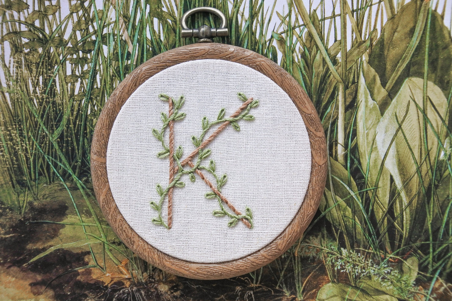 Botanical letter K embroidery hoop with eucalyptus green vines and pale brown stitched letter on neutral fabric