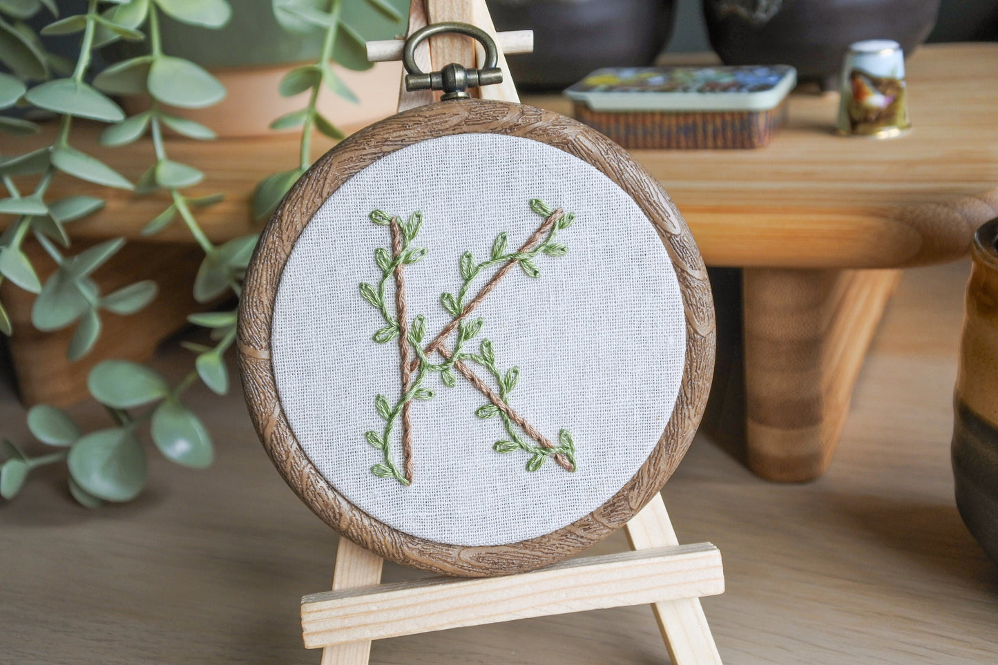 Botanical letter K embroidery hoop with eucalyptus green vines and pale brown stitched letter displayed on a wooden easel against a soft background