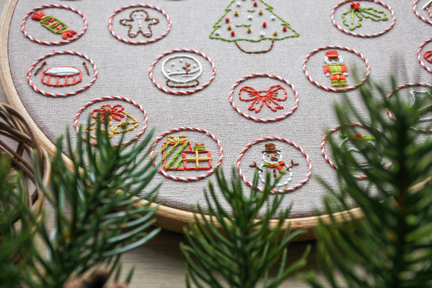 Closeup of hand embroidered Christmas advent calendar design showing festive stitched details, including a Christmas tree and mini holiday motifs. Pine branches blurred in the foreground.