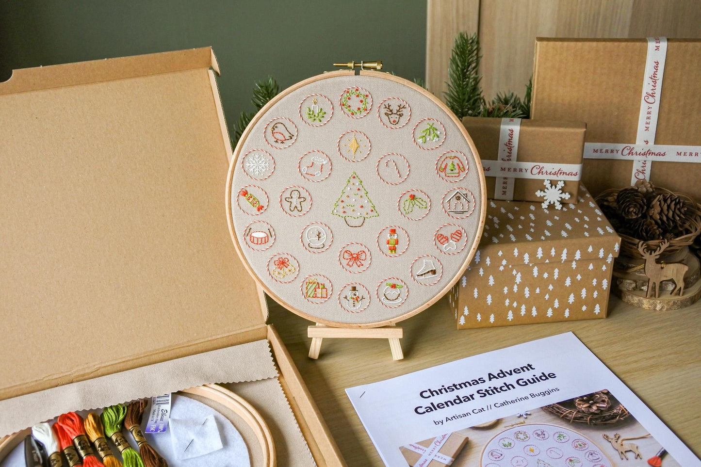 Hand embroidered Christmas advent calendar hoop displayed on a mini wooden easel, surrounded by stitch guide and kit box with embroidery materials.
