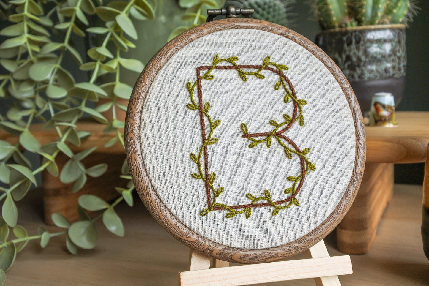 Details of hand embroidery with jungle green leafy vines wrapped around the dark brown letter B. Stitched on light neutral fabric in wood effect hoop frame.