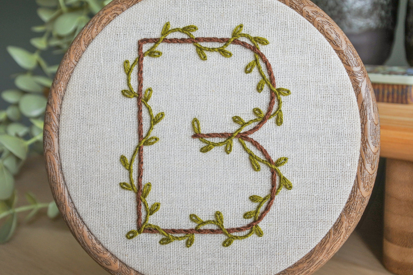 Closeup of hand-embroidered botanical letter 'B' hoop, featuring jungle green leafy vines and dark brown thread on light neutral fabric.