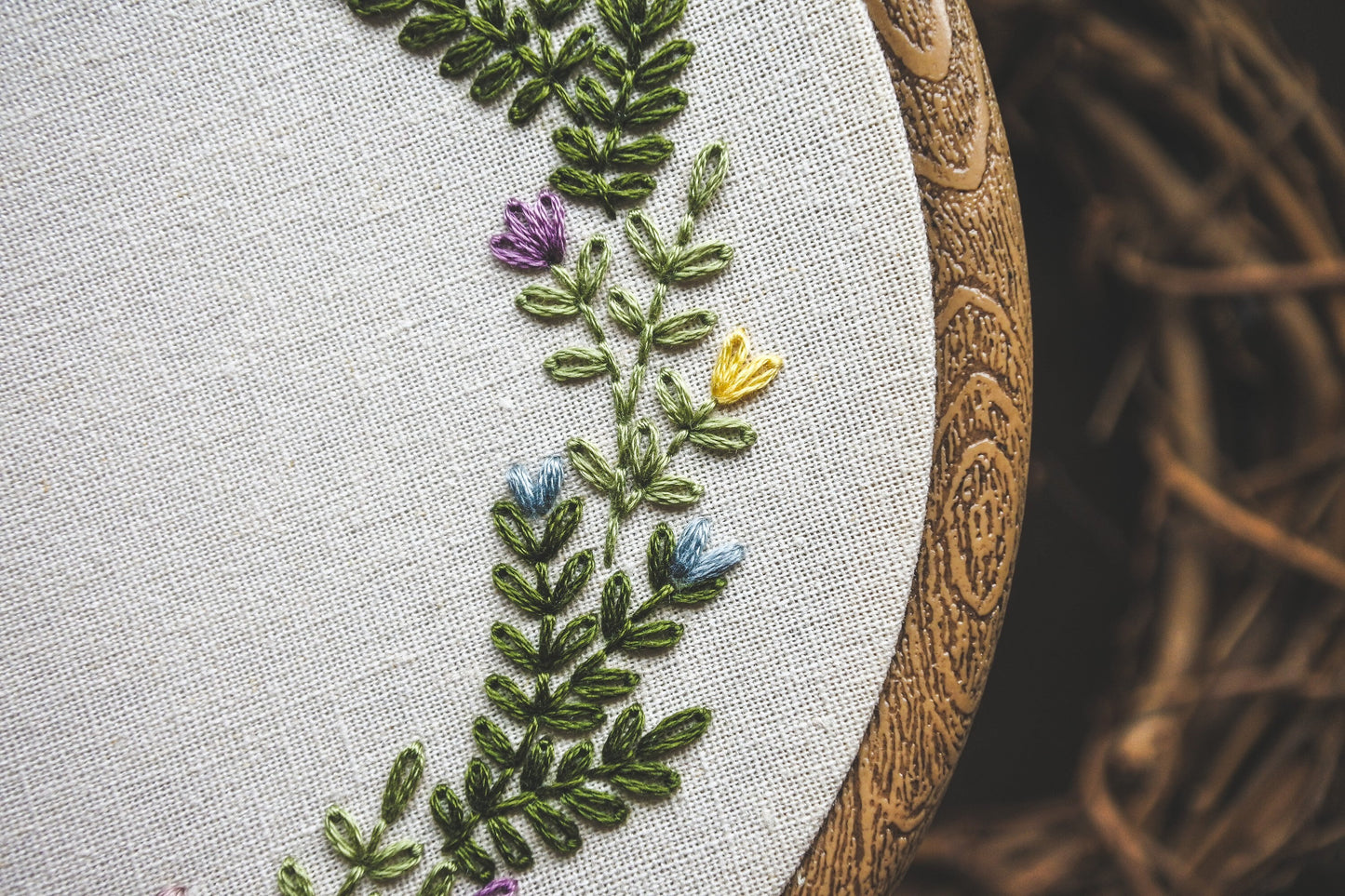 Close-up of hand embroidered spring flowers and green foliage. The flowers are yellow, purple and blue. Also shows detail of wood effect embroidery hoop.