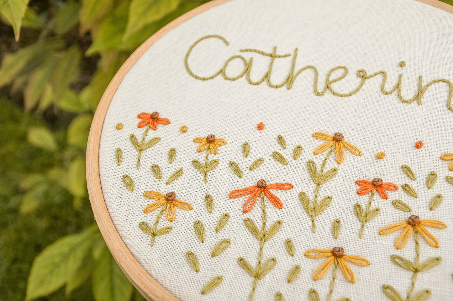 Close-up of the mustard yellow and warm orange coneflowers, with warm green stems and leaves, on the personalised embroidery hoop