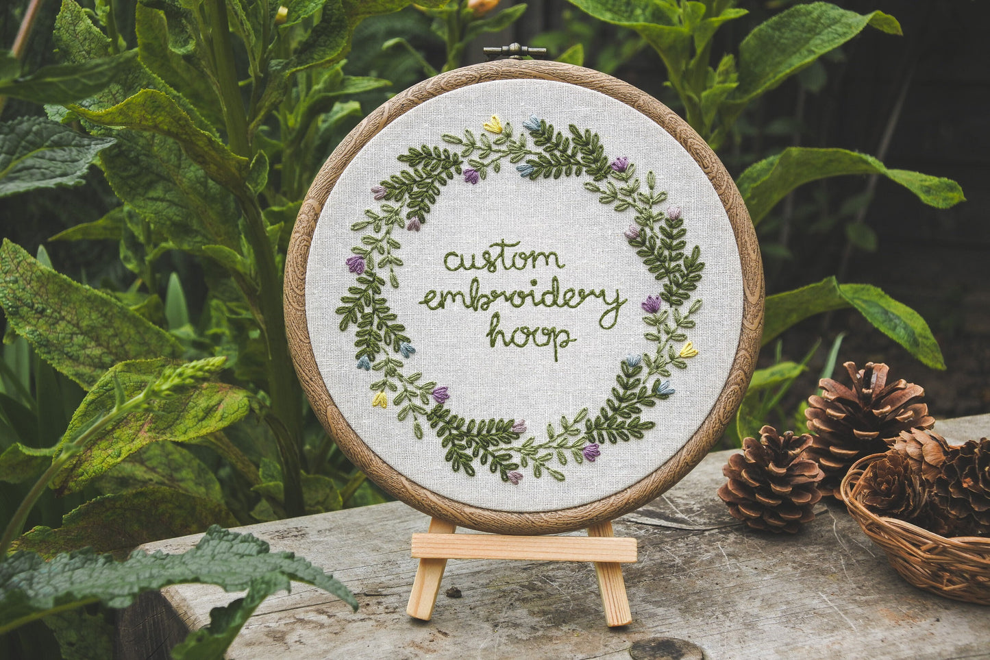 Custom Spring floral embroidery hoop displayed on a mini wooden easel outside, surrounded by greenery and pinecones.