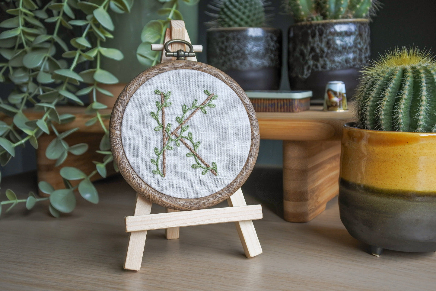 Botanical letter K embroidery hoop with eucalyptus green vines and pale brown letter displayed on a small wooden easel