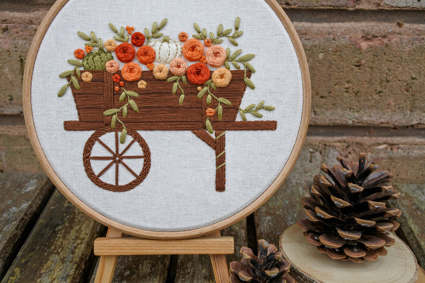 Autumn floral pumpkin wagon embroidery displayed in a wooden hoop on a mini wooden easel next to pinecones