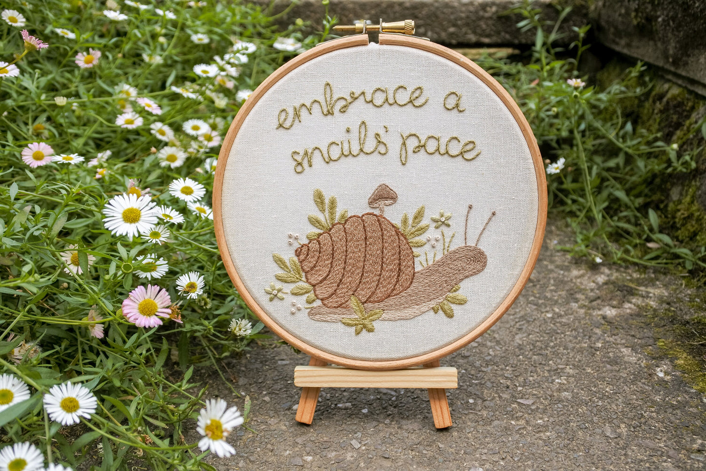 The finished embroidery displayed outside on a mini wooden easel surrounded by daisy plants