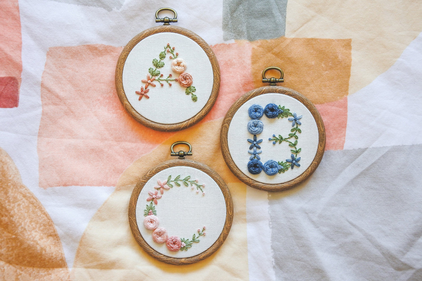 Hand-embroidered floral letter A, B, and C hoops featuring peach, blue, and pink flowers with greenery.
