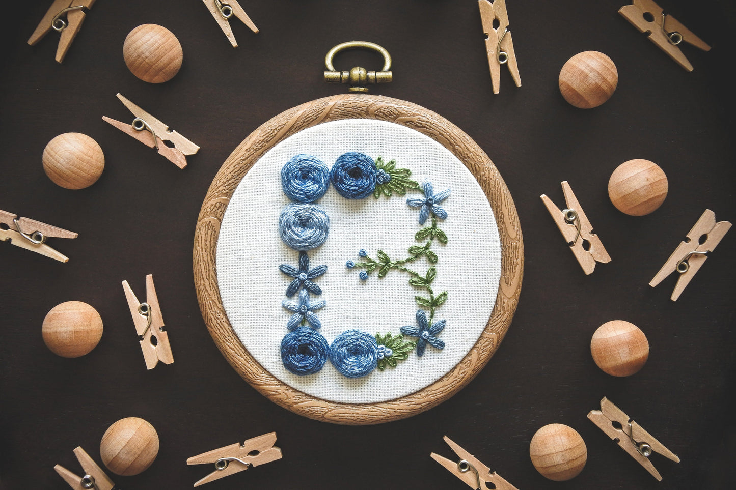 Hand-embroidered blue floral letter 'B' hoop with green foliage. Decorative background of mini wooden pegs and balls.