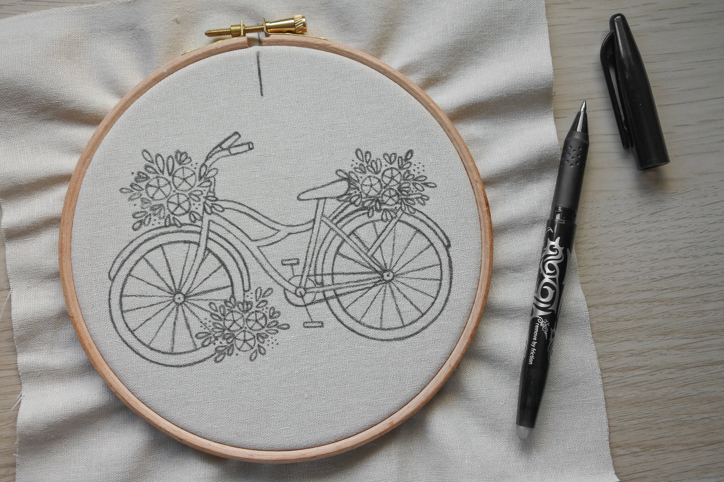 Floral bicycle embroidery pattern transferred onto neutral cotton fabric with a heat erasable pen placed beside the hoop.