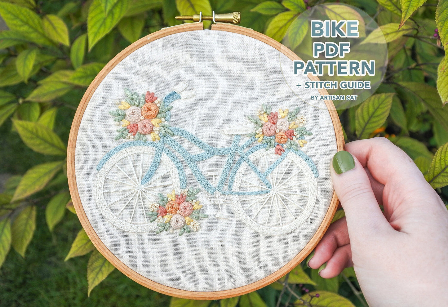 Hand holding finished floral bicycle embroidery with pastel flowers, featuring text "Floral Bike PDF Pattern and Stitch Guide by Artisan Cat. Background is green leaves out of focus.