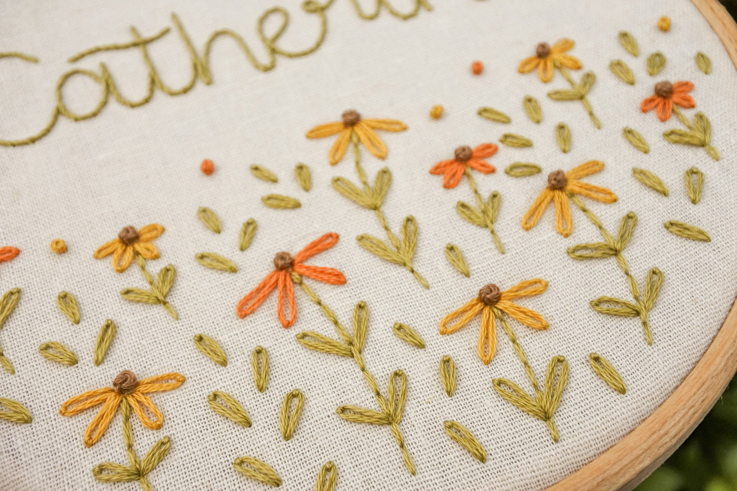 Close-up of the personalised coneflower name embroidery hoop, showcasing the name Catherine above warm orange and mustard yellow coneflowers and green stems.