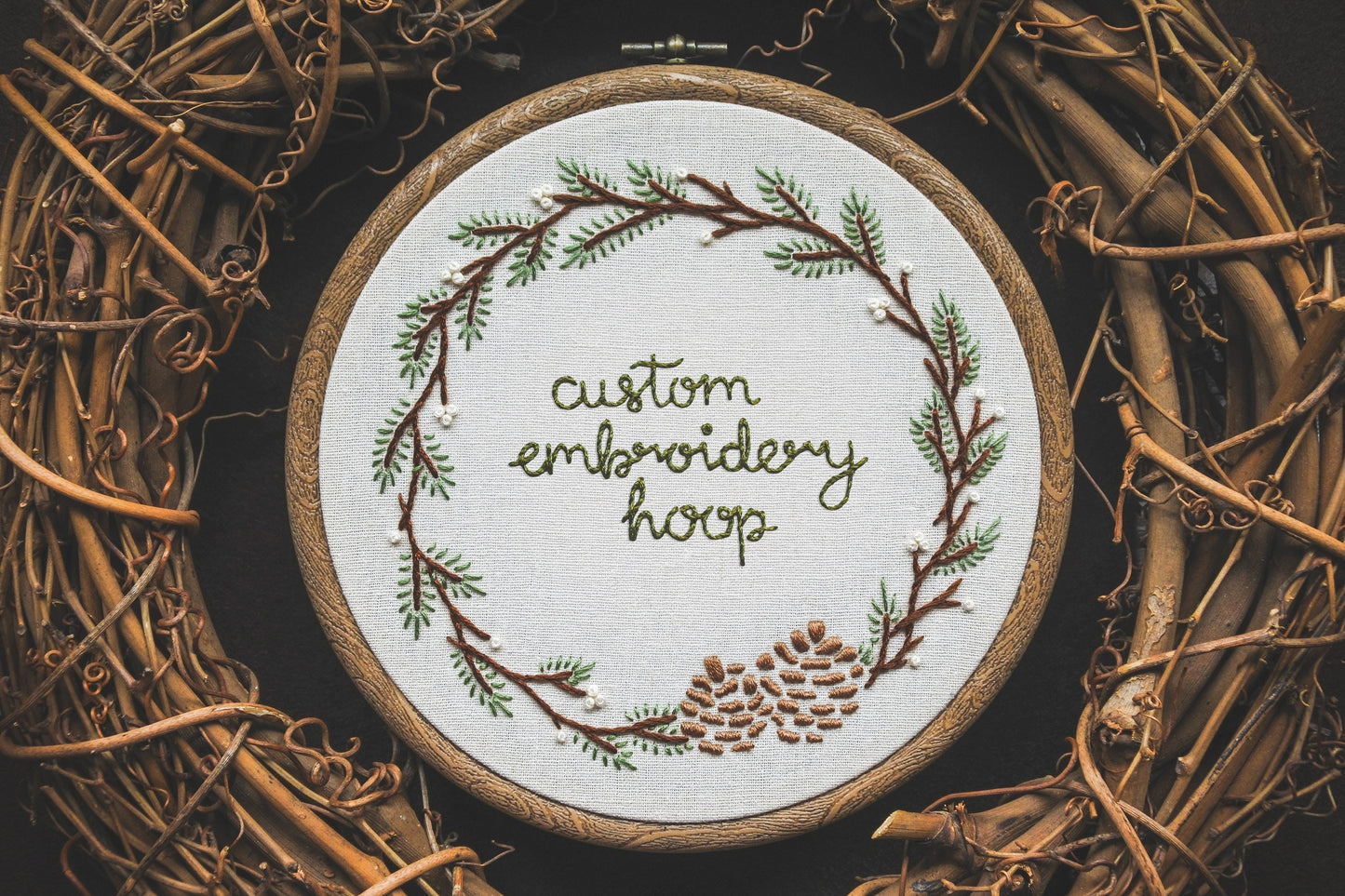 Hand-embroidered winter wreath hoop with pinecones, conifer branches, and white berries. 'Custom embroidery hoop' lettering in the centre.