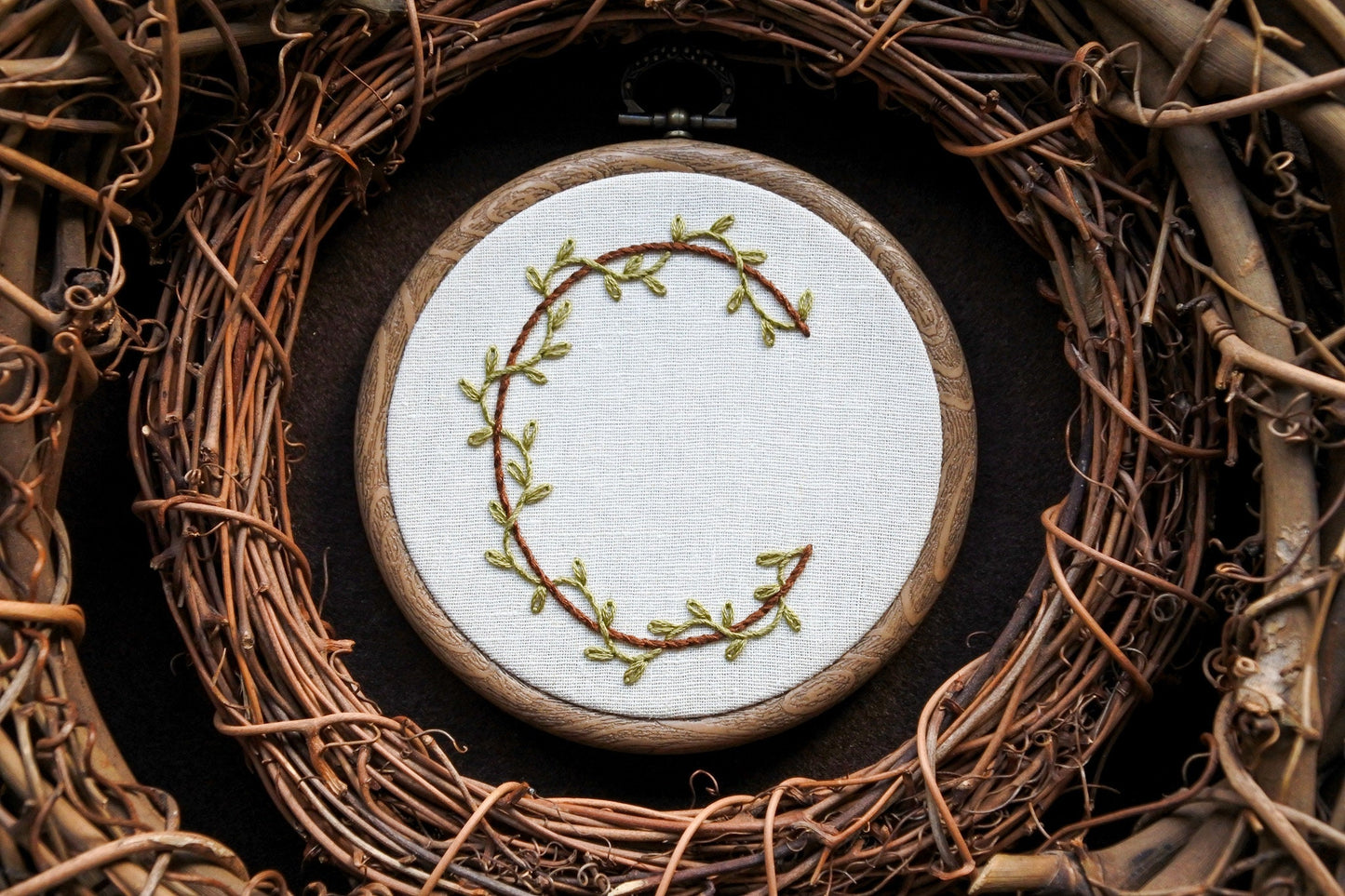 Hand embroidery of botanical letter C with green leafy vines and brown stitched initial in a wood-effect hoop