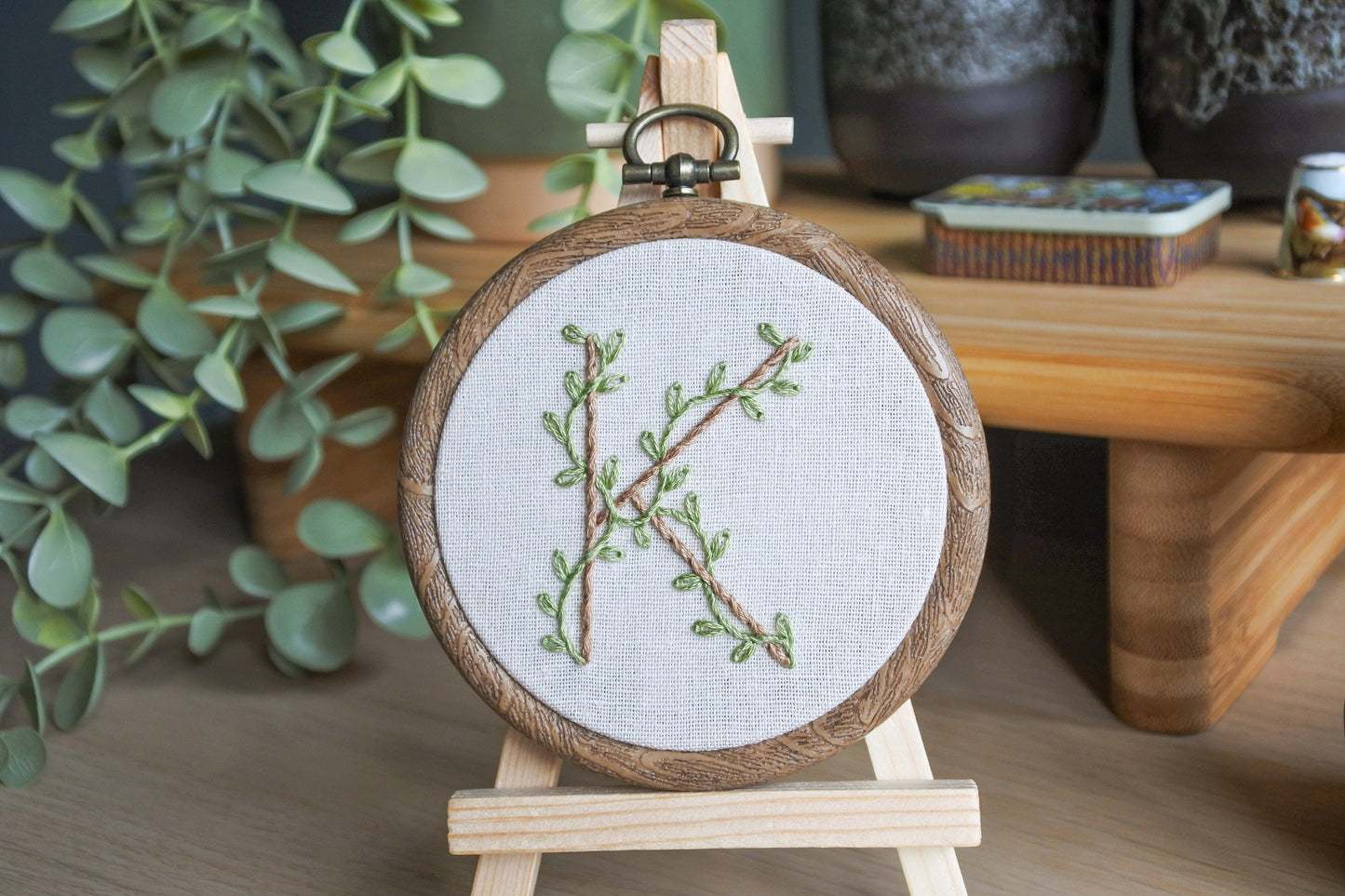 Hand embroidery of botanical letter K with eucalyptus green vines and pale brown stitched letter in a 3-inch wood-effect hoop