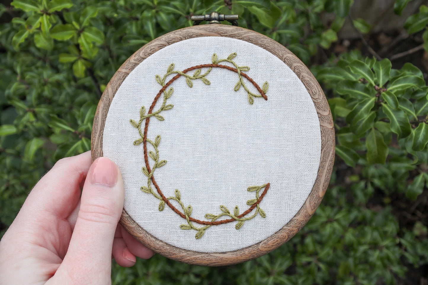 Hand holding a botanical letter C embroidery hoop with green vines and brown stitched letter on neutral fabric