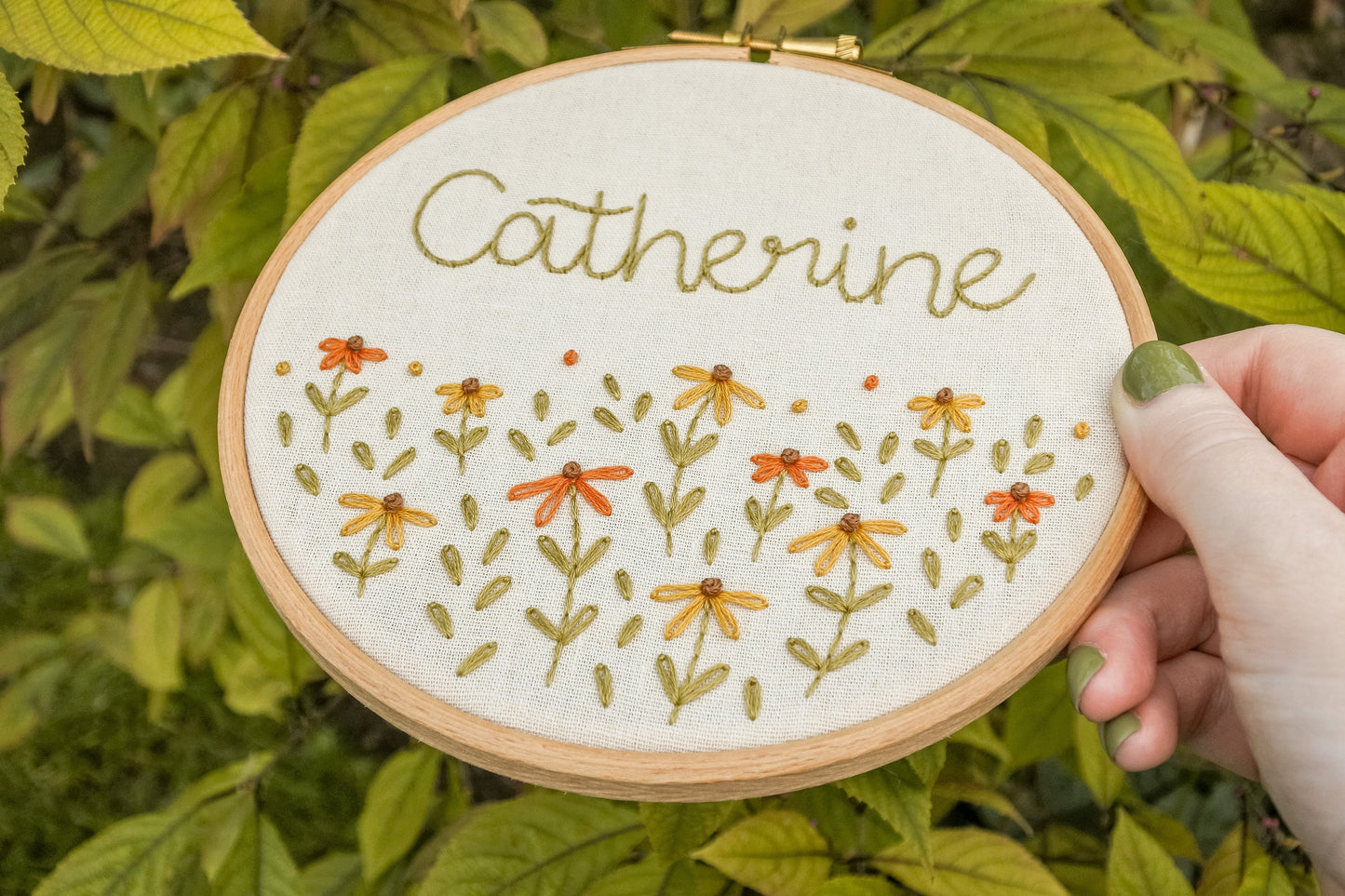 Hand holding the personalised coneflower name embroidery hoop at an angle, featuring the name Catherine and coneflowers in warm orange and mustard yellow.