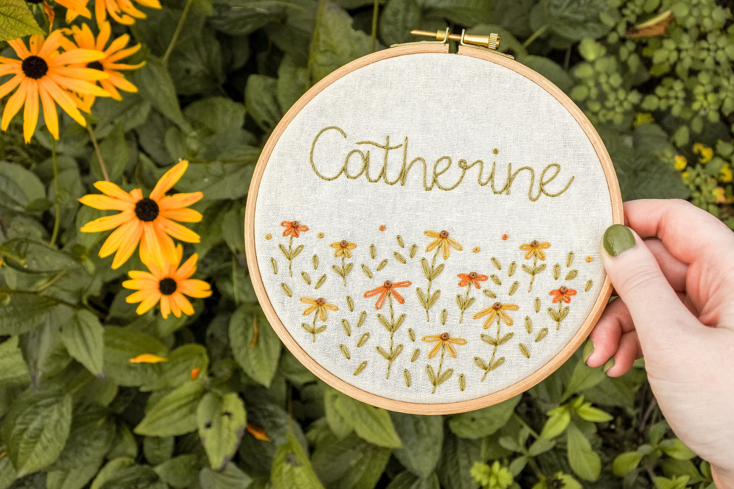 Hand holding the personalised coneflower name embroidery hoop at an angle, with warm orange and mustard yellow coneflowers and warm green leaves.