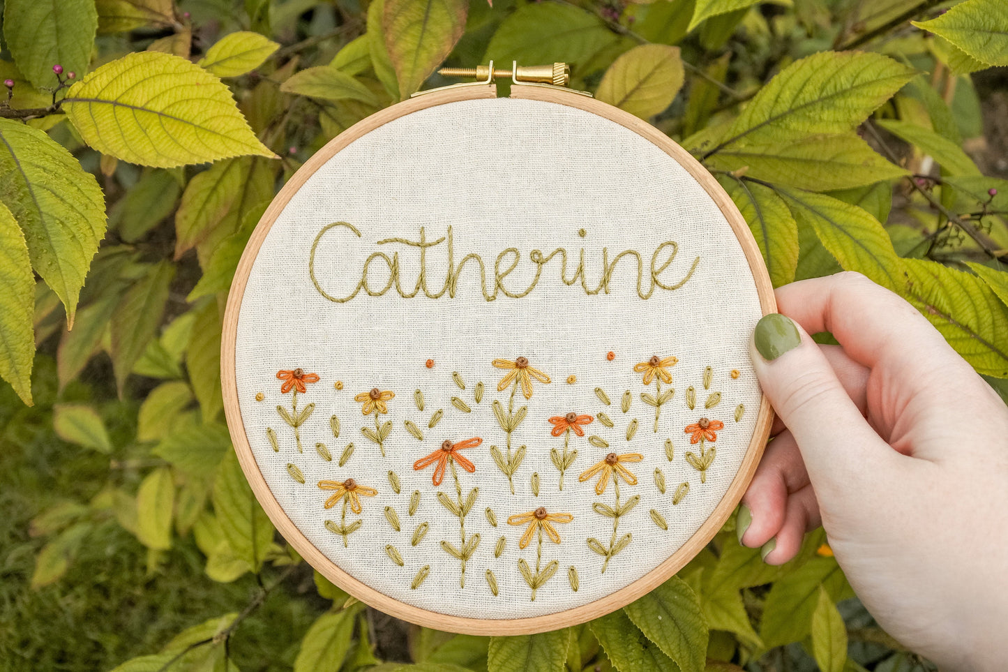 Hand holding the personalised coneflower name embroidery hoop featuring the name Catherine, surrounded by warm green foliage in the background.