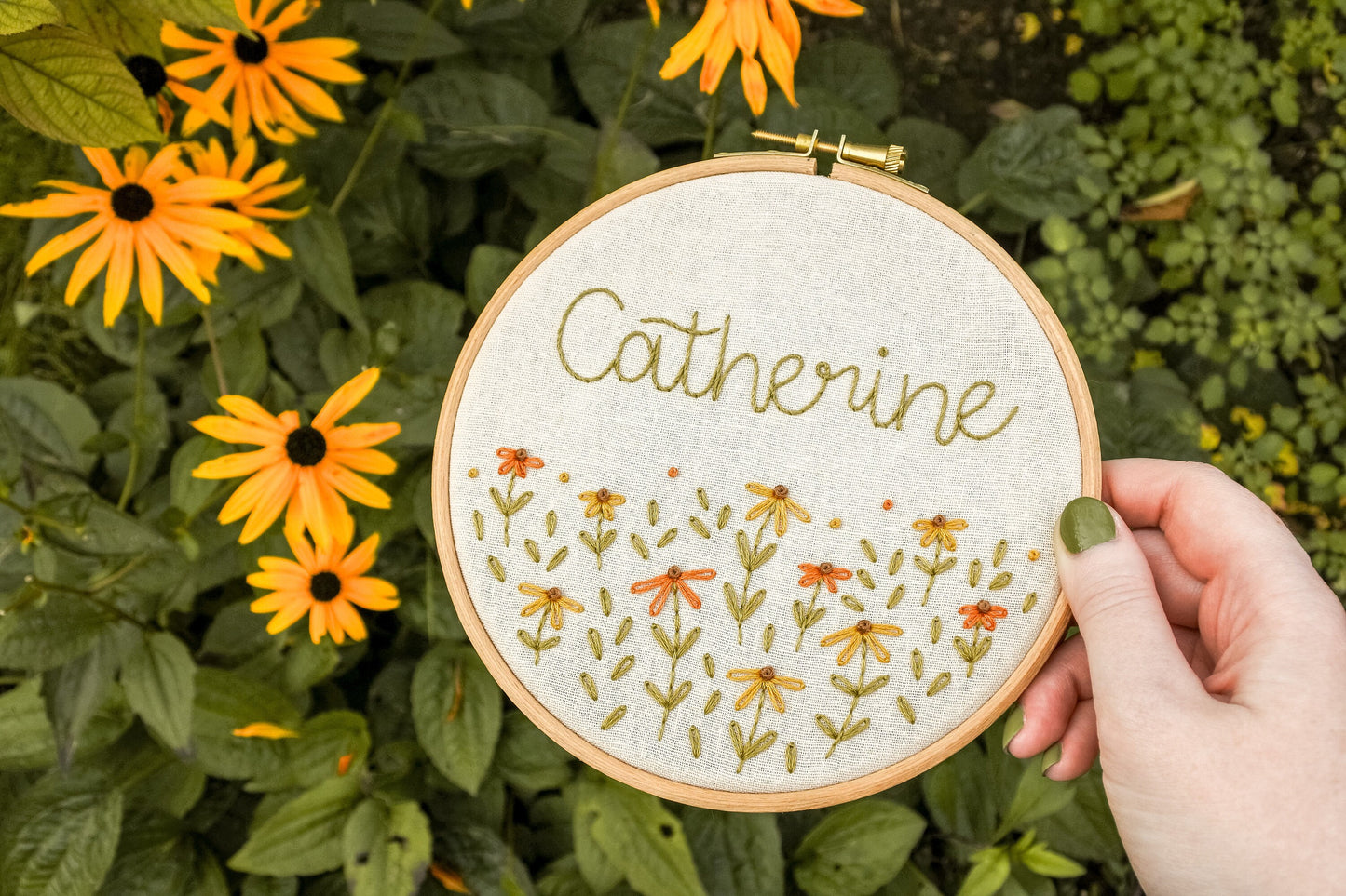 Hand holding the personalised coneflower name embroidery hoop next to yellow Rudbeckia coneflowers, featuring the name Catherine above the flowers.