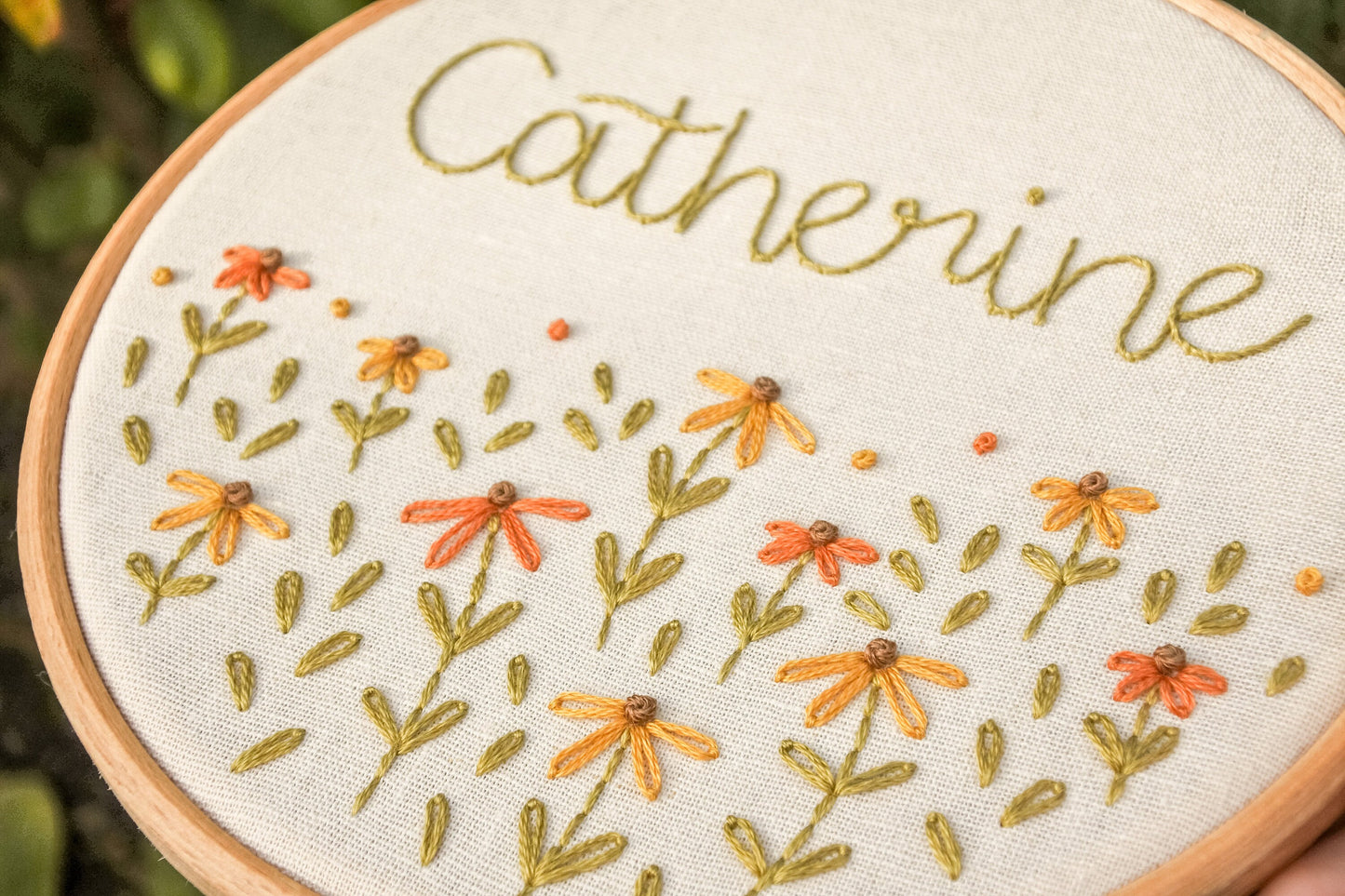 Hand holding the personalised coneflower name embroidery hoop at an angle, featuring the name Catherine with coneflowers in warm tones and green leaves.