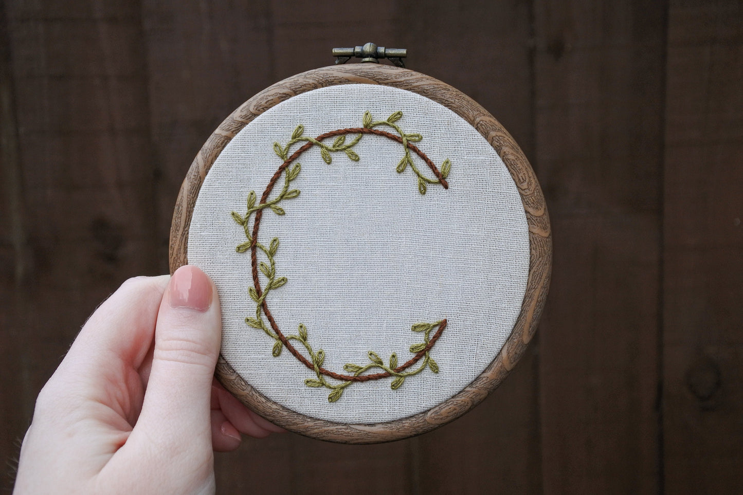 Hand holding botanical letter C embroidery hoop showing green vine and brown letter details on neutral fabric