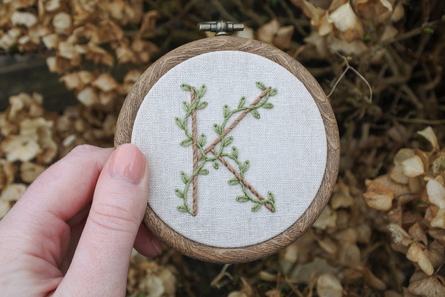 Hand holding botanical letter K embroidery hoop with eucalyptus green vines and pale brown stitched letter on neutral fabric