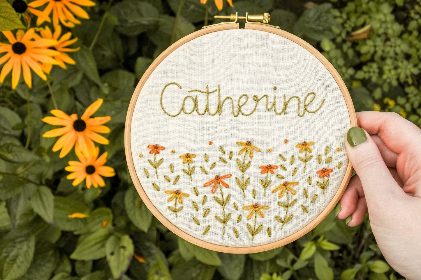 Hand holding the personalised coneflower name embroidery hoop featuring the name Catherine above warm orange and mustard yellow coneflowers, with warm green stems and leaves.