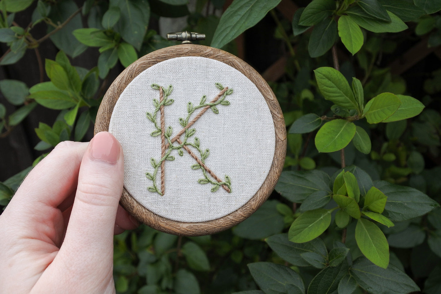 Hand holding letter k embroidery hoop art, featuring pale brown hand stitched letter with eucalyptus green leafy vines wrapping around it