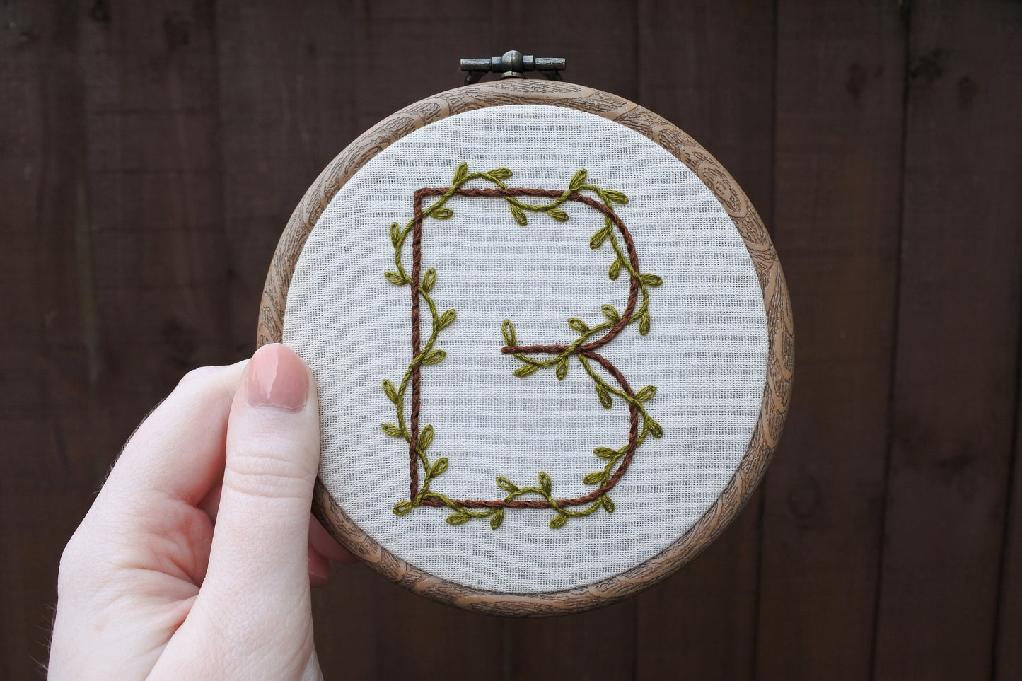 hand holding botanical embroidered letter hoop art featuring jungle green leafy vines around dark brown thread
