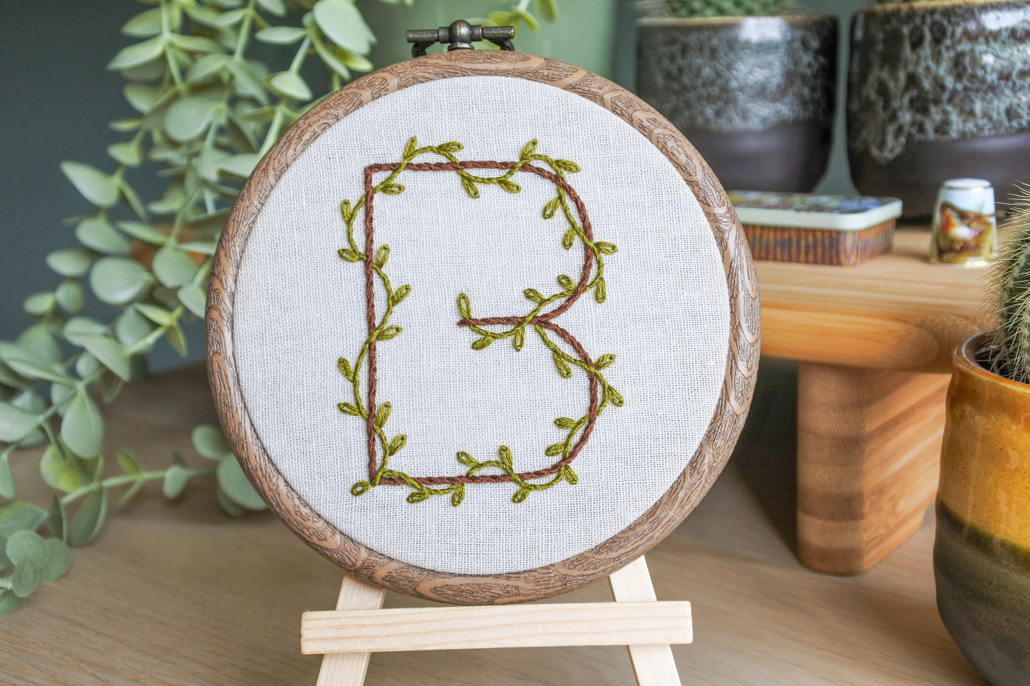 hand stitched botanical letter b embroidery hoop art on a wooden easel. The letter b is stitched with dark brown thread with leafy green vines wrapping around it.